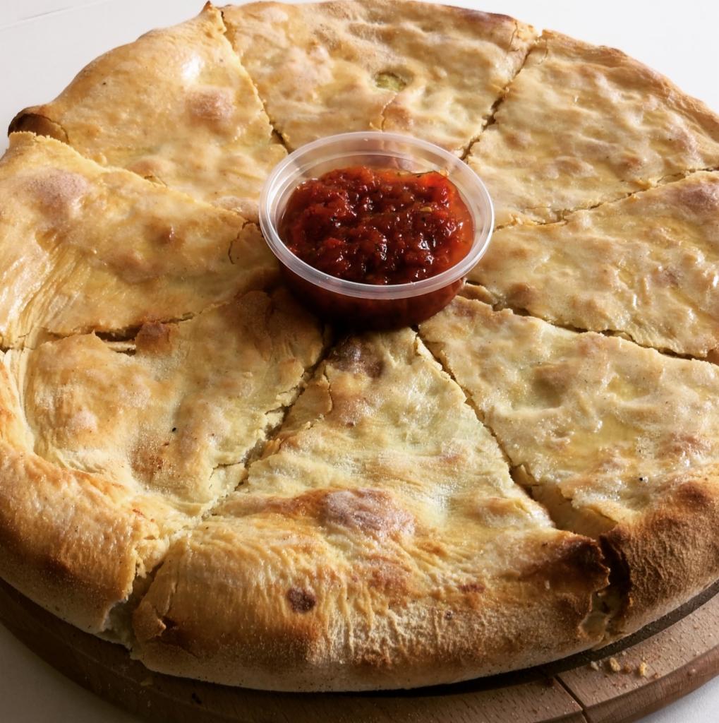 Okrągły placek podzielony na trójkątne porcje na drewnianej desce, z sosem pomidorowym w plastikowym pojemniku pośrodku.