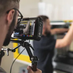 Operator kamery z ekranem podglądowym filmuje pracownika obsługującego maszynę przemysłową w hali produkcyjnej.