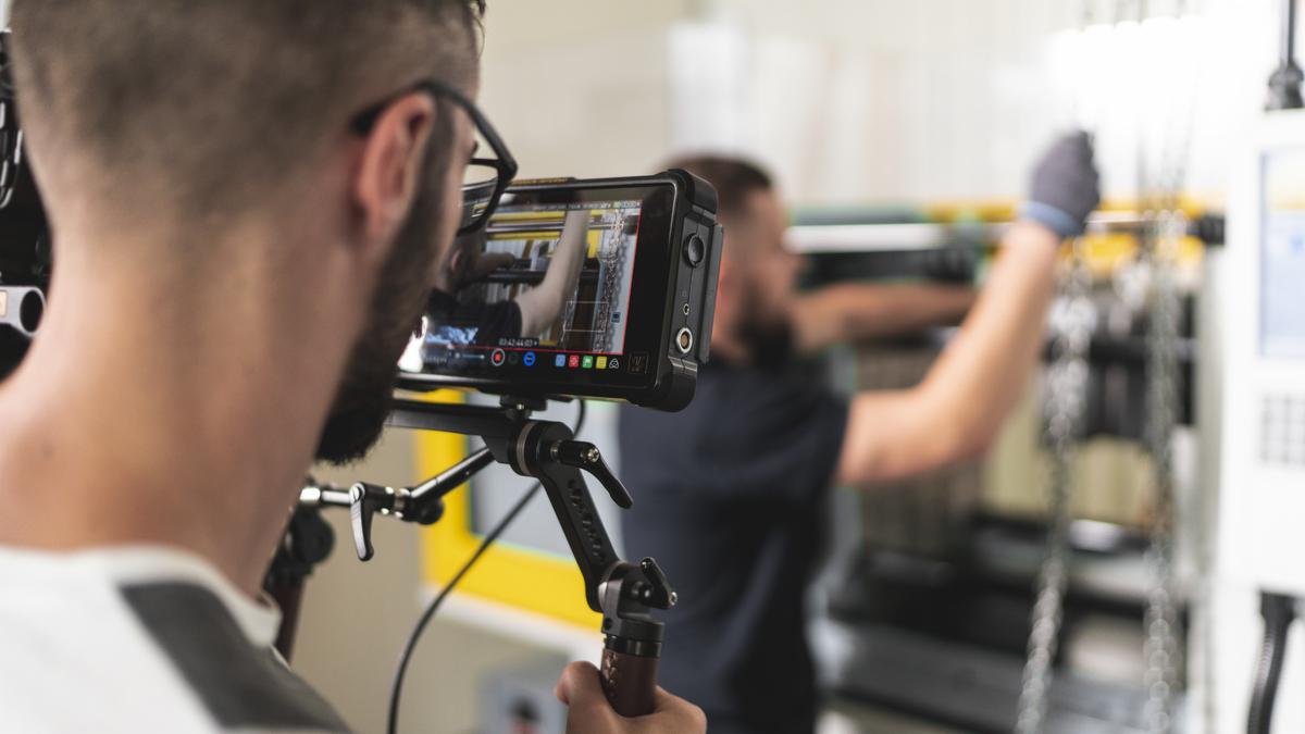 Operator kamery z ekranem podglądowym filmuje pracownika obsługującego maszynę przemysłową w hali produkcyjnej.