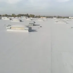 Jasnoszary, płaski dach przemysłowy z licznymi świetlikami i odpowietrznikami, widok z góry na tle zabudowań miejskich.