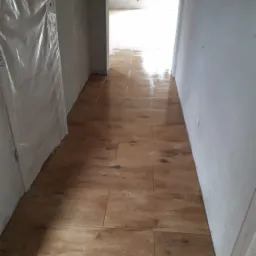 Położona, błyszcząca podłoga z płytek imitujących drewno w wąskim korytarzu podczas remontu. Drzwi po lewej stronie zabezpieczone folią ochronną, ściany w trakcie malowania na biało.