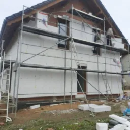 Dwukondygnacyjny dom w trakcie ocieplania styropianem, widoczny fragment elewacji z zamontowanymi płytami, rusztowanie dookoła budynku i elementy styropianu na ziemi.