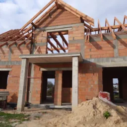 Surowy stan domu w trakcie budowy, z widoczną konstrukcją dachu z drewnianych belek i ścianami z czerwonej cegły, na pierwszym planie pryzma piasku.