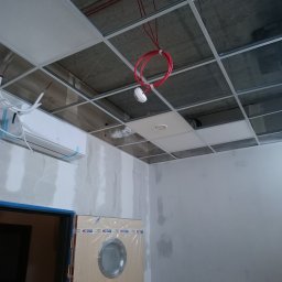 Startklima - Wnętrze w trakcie prac wykończeniowych: widoczny klimatyzator na ścianie, częściowo zamontowany sufit podwieszany z widocznymi przewodami i czujnikiem dymu, fragment drzwi z niebieską ramą...