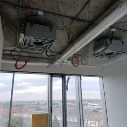 Dwa szare moduły wentylacyjne zamontowane pod surowym betonowym stropem w budynku komercyjnym z widocznymi przewodami i aluminiowymi rurami wentylacyjnymi, widok przez okno na zabudowę miejską.