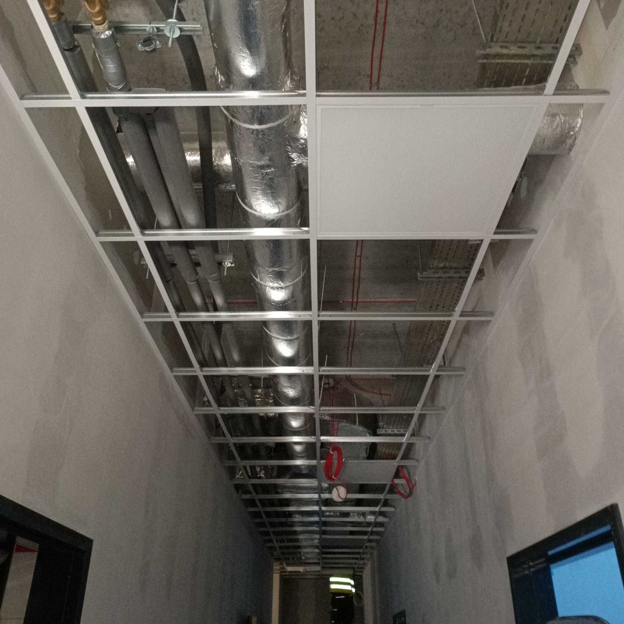Widok z dołu na sufit podwieszany w trakcie montażu we Wrocławiu, widoczne rury, kable i kanały wentylacyjne, częściowo zakryte płytami sufitowymi.