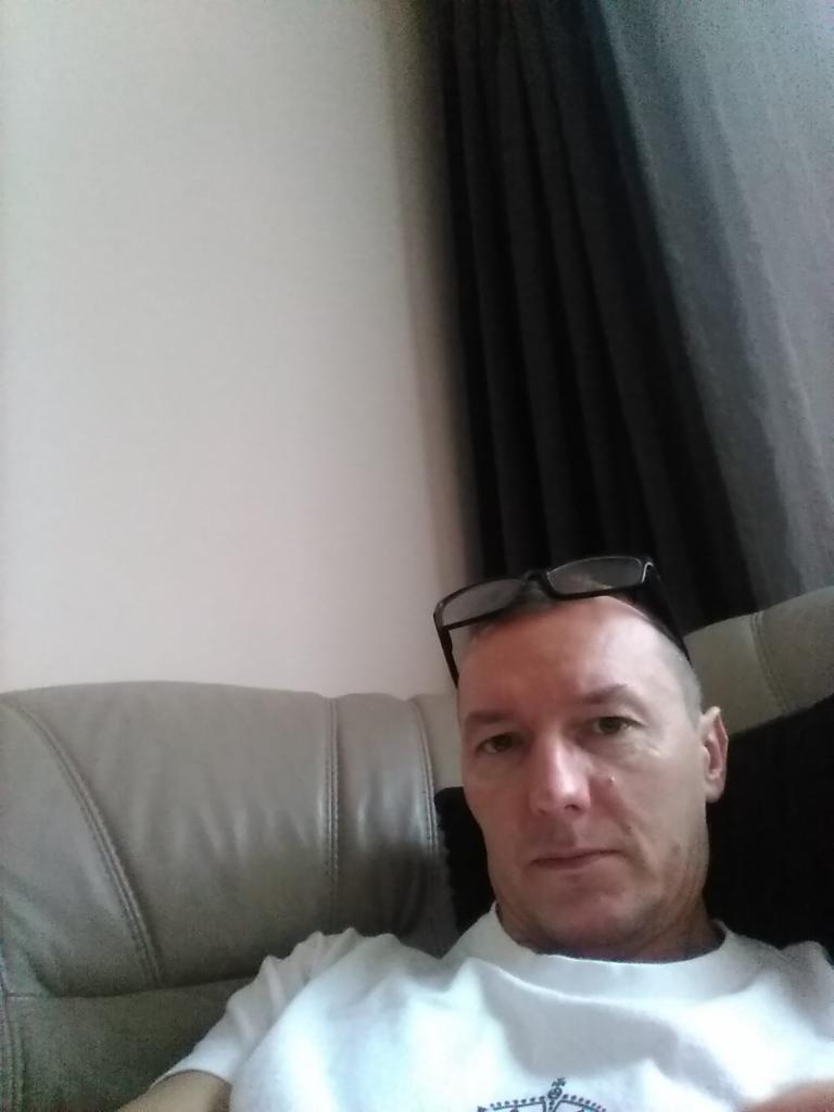 Portret mężczyzny w białej koszulce, z okularami na głowie, siedzącego na skórzanym fotelu na tle ściany i zasłon.