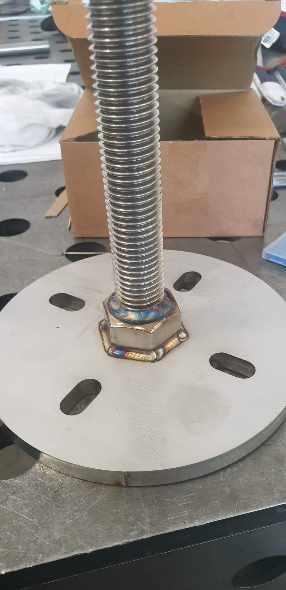 Metalowa śruba z nakrętką, przyspawana do okrągłej podstawy z otworami, widoczne ślady spawania z tęczowymi przebarwieniami, wykonane w Katowicach.