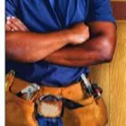 Mężczyzna w niebieskiej koszulce z założonymi rękami, ubrany w skórzany pas monterski z narzędziami, stojący na tle drewnianej ściany. Usługi w Tarnowie.