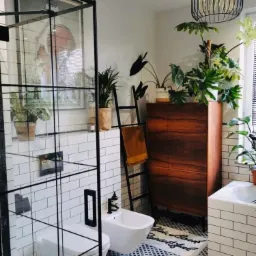 Nowoczesna łazienka z kabiną prysznicową typu walk-in z czarnymi profilami, wiszącą toaletą i bidetem, czarno-białą mozaiką na podłodze, drewnianą komodą z roślinami doniczkowymi i dekoracyjną...