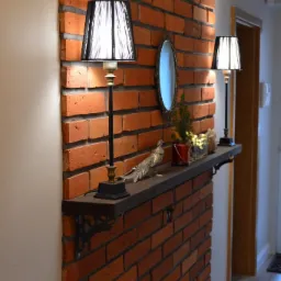 Ściana z czerwonej cegły z drewnianą półką, na której stoją dwie lampy z czarnymi abażurami, srebrna figurka ptaka i małe lusterko w ozdobnej ramie.