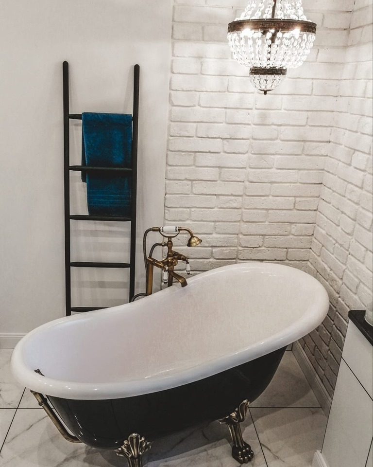 Elegancka łazienka z wolnostojącą wanną na lwich nóżkach, baterią w stylu retro i dekoracyjną drabinką na ręcznik, oświetlona kryształowym żyrandolem.