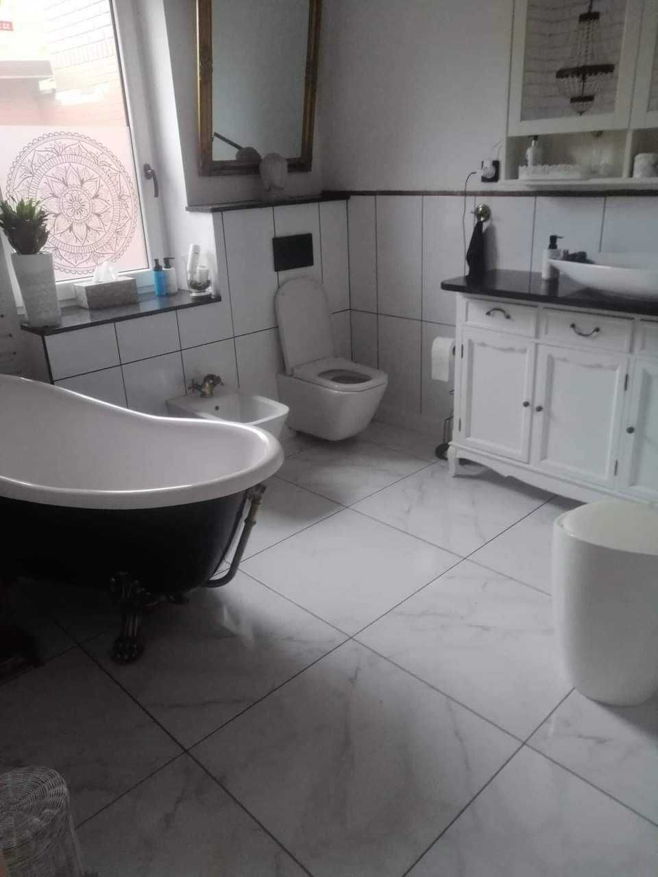 Elegancka łazienka w stylu retro z wolnostojącą wanną na ozdobnych nóżkach, bidetem, toaletą, umywalką z szafką i dekoracyjnym lustrem w złotej ramie, uzupełniona o białe płytki i dekoracje okienne.