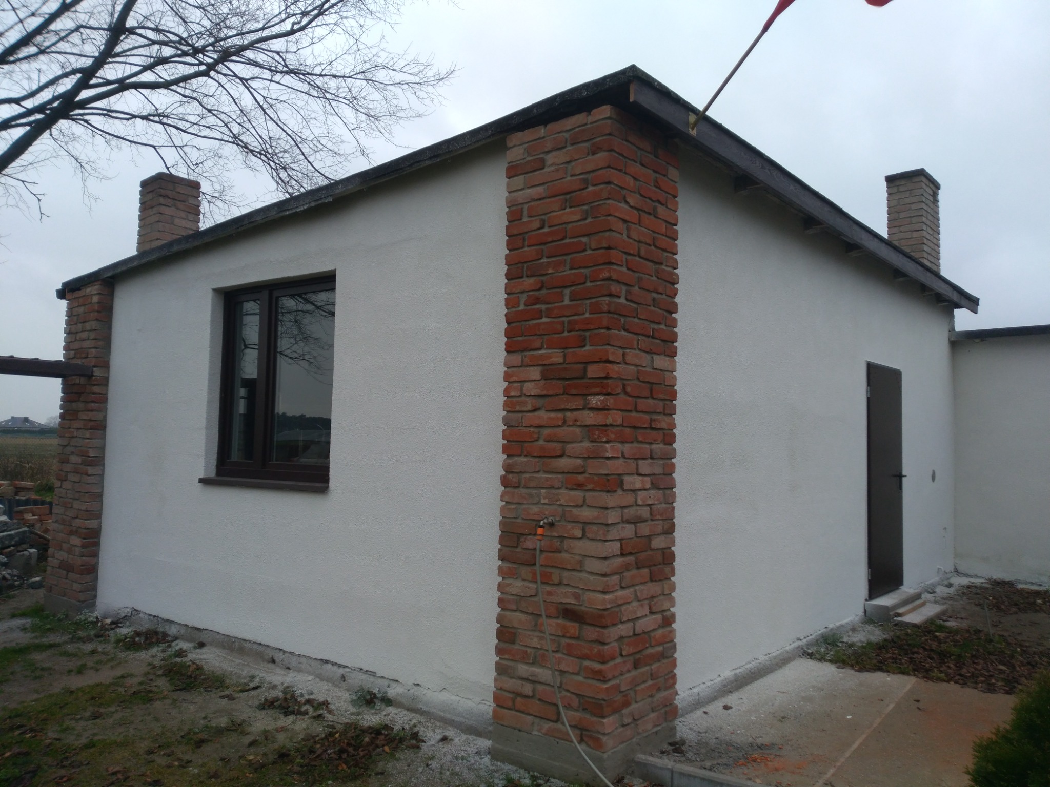 Otynkowany budynek z ceglanymi elementami dekoracyjnymi, widoczna elewacja z oknem i drzwiami wejściowymi, fragment ogrodu z resztkami liści.