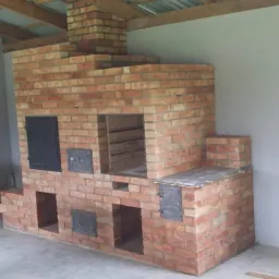 Murowany piec zewnętrzny z cegły, z czarnymi, metalowymi drzwiczkami i otwartymi wnękami, pod zadaszeniem z drewnianą konstrukcją.