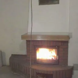 Murowany kominek z cegły z palącym się ogniem, drewniana półka nad kominkiem, kratka wentylacyjna na ścianie, surowy stan wykończenia wnętrza.