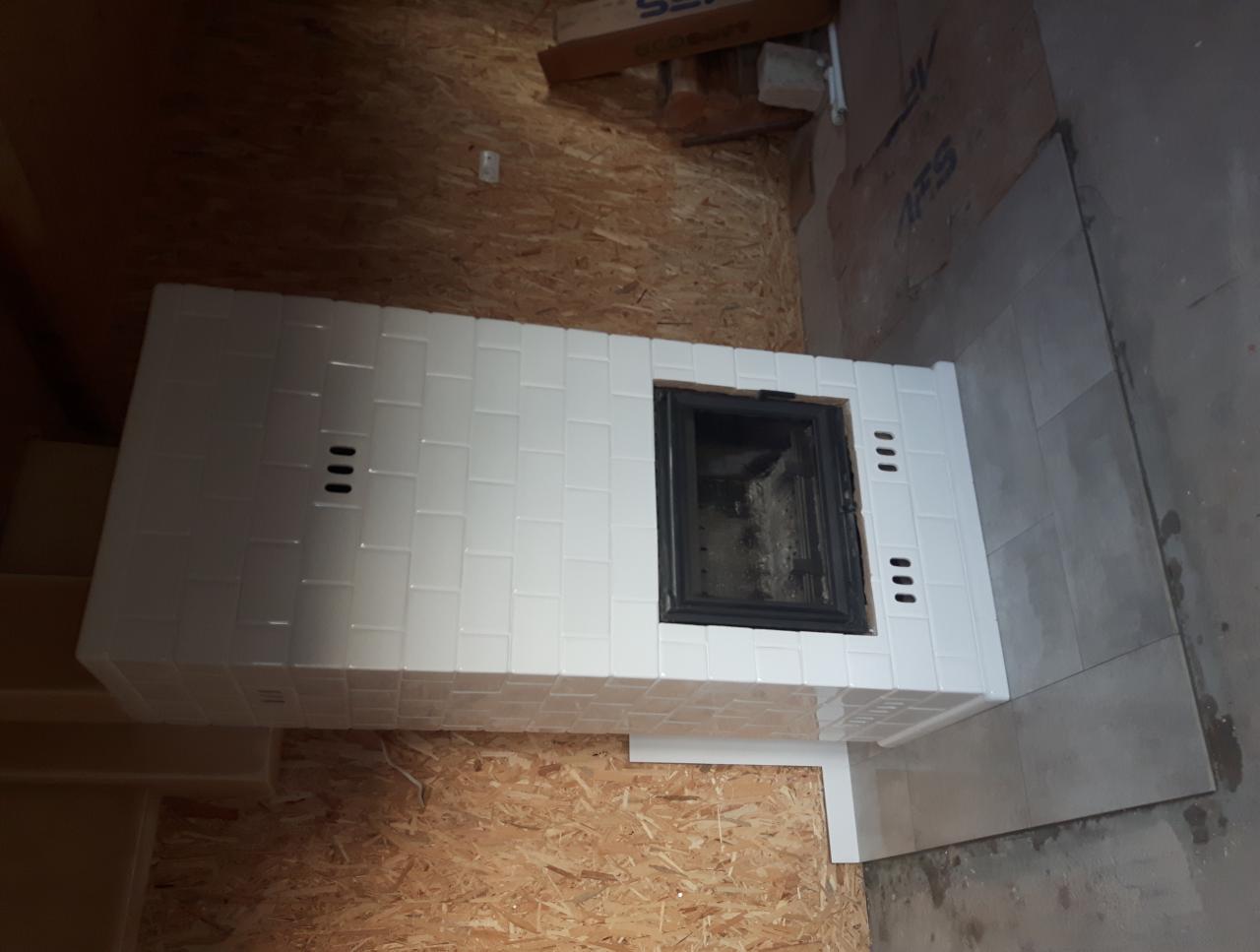 Wykończony piec kaflowy z białymi płytkami, widoczne trzy rzędy otworów wentylacyjnych, czarne drzwiczki paleniska, obok ułożone szare płytki podłogowe, surowa ściana z płyty OSB w tle.