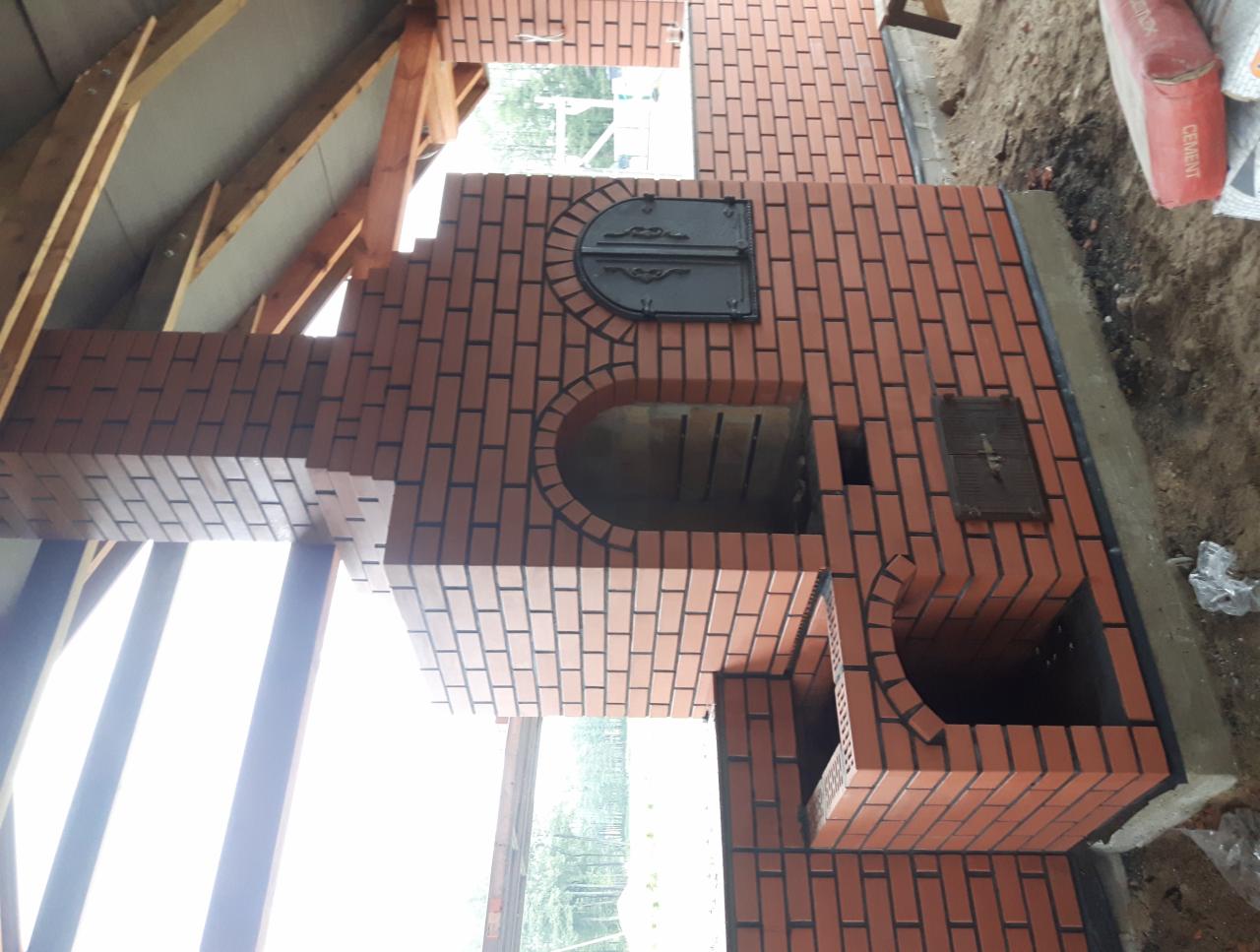 Murowany grill z czerwonej cegły z żeliwnymi drzwiczkami, widok na konstrukcję z drewna i cement w worku obok.