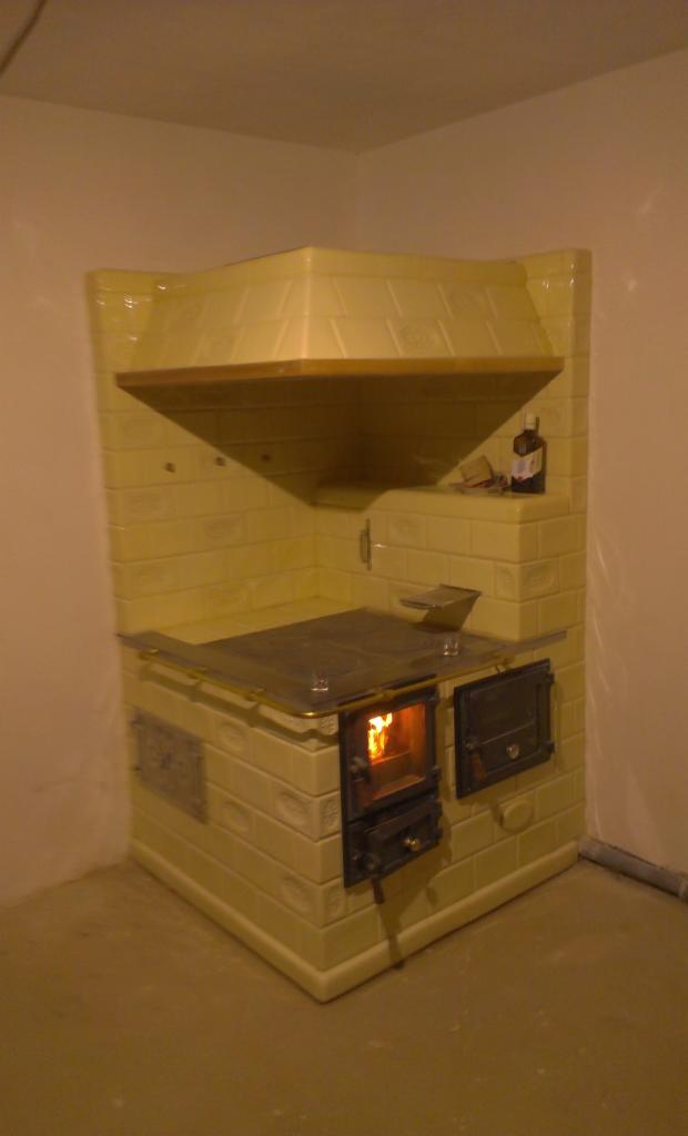 Żółty, kaflowy piec narożny z paleniskiem, na którym widać płomienie, w rogu pomieszczenia z beżowymi ścianami i podłogą.