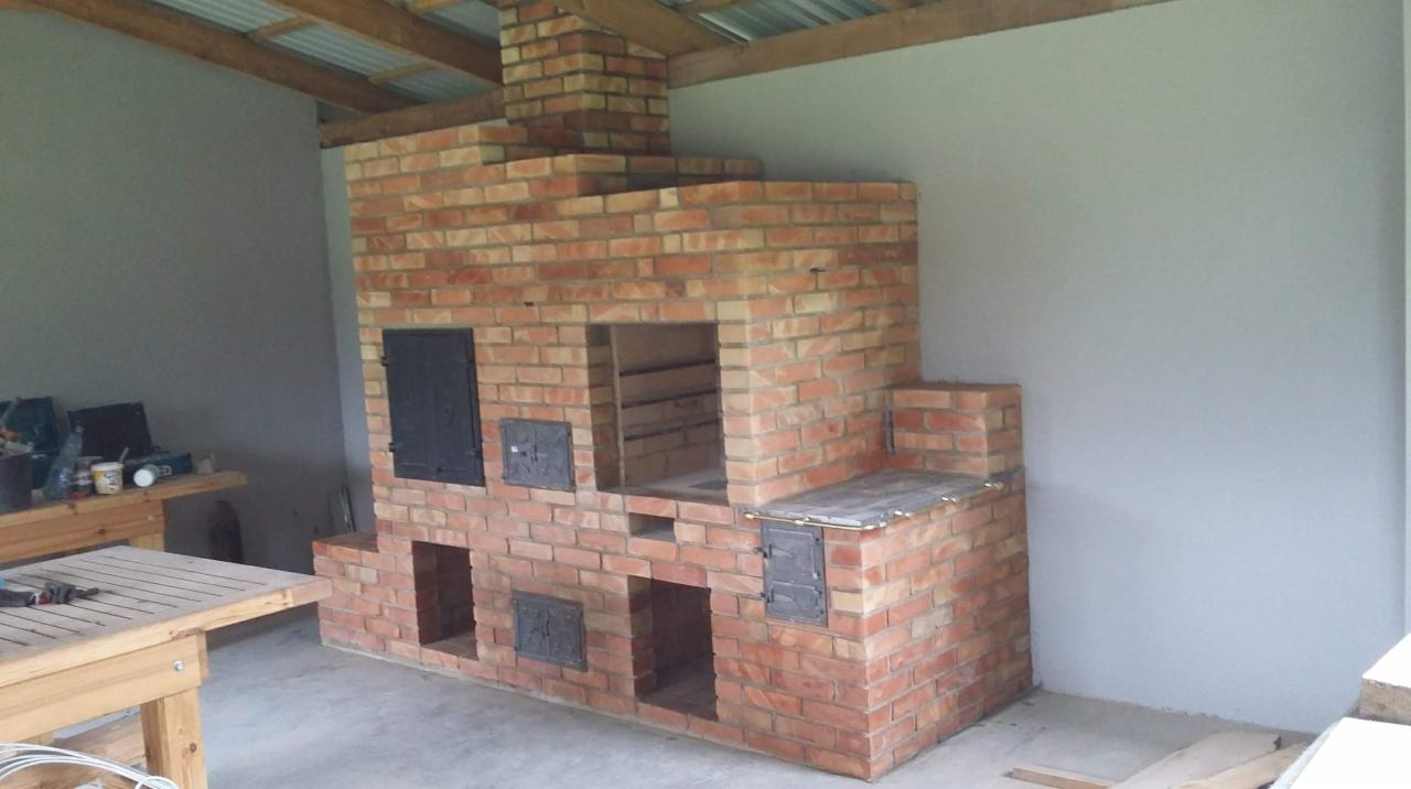 Murowany piec zewnętrzny z cegły, z czarnymi, metalowymi drzwiczkami i otwartymi wnękami, pod zadaszeniem z drewnianą konstrukcją.