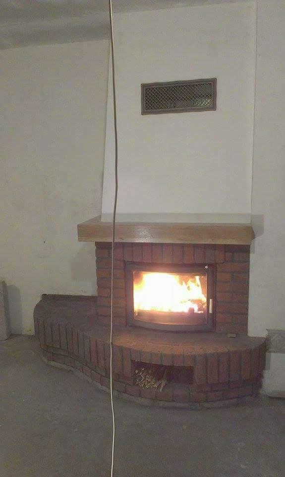 Murowany kominek z cegły z palącym się ogniem, drewniana półka nad kominkiem, kratka wentylacyjna na ścianie, surowy stan wykończenia wnętrza.