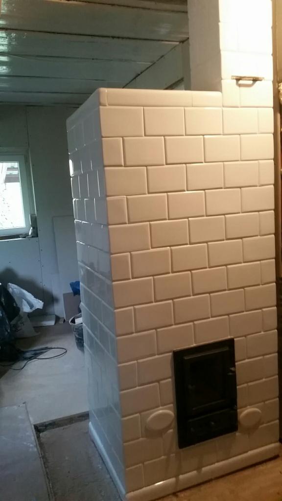 Biały piec kaflowy z czarnymi drzwiczkami w trakcie budowy lub renowacji, widoczne niedokończone wykończenia podłogi i sufitu.