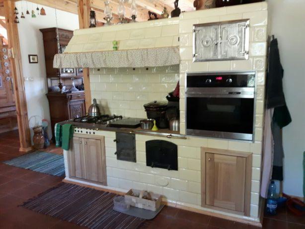 Kuchenna zabudowa z piecem kaflowym, piekarnikiem i płytą grzewczą, wykończona jasnymi, kwadratowymi kaflami z delikatnym wzorem, widoczna w rustykalnym wnętrzu z drewnianymi elementami.