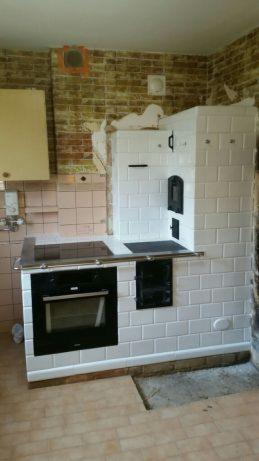 Biały piec kaflowy z piekarnikiem i płytą grzewczą w kuchni w trakcie remontu, widoczne odsłonięte cegły i brakujące płytki podłogowe.