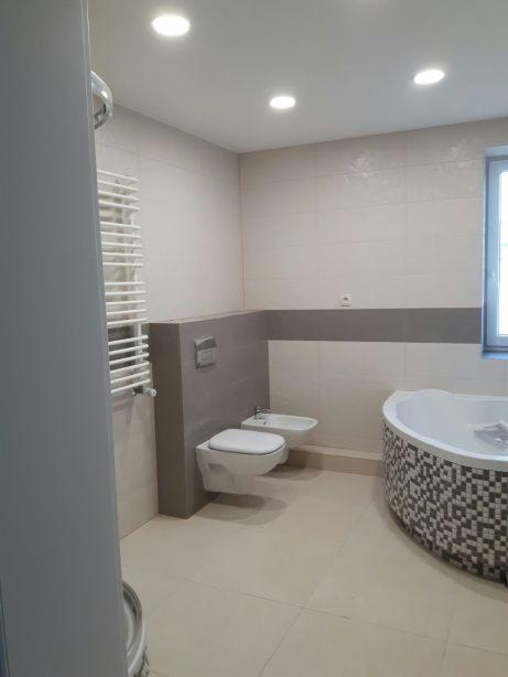 Jasna łazienka po remoncie z beżowymi płytkami, wiszącą toaletą i bidetem oraz wanną obudowaną mozaiką w odcieniach szarości i bieli, widoczny grzejnik drabinkowy.