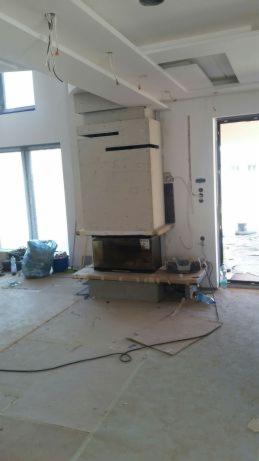 Wnętrze w trakcie remontu z widocznym kominkiem obłożonym płytami gipsowo-kartonowymi przed szpachlowaniem, widoczne kable i worki z materiałami budowlanymi.