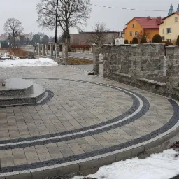 Ułożona kostka brukowa przed domem z elementami dekoracyjnymi w postaci ciemniejszych pas&oacute;w i murk&oacute;w z kamienia, częściowo pokryta śniegiem.