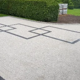 Wyłożona kostką brukową powierzchnia z dekoracyjnym wzorem z ciemnej kostki tworzącym geometryczne figury, obrzeża trawników i rabat również wykończone kostką, w tle zielony żywopłot i trawnik.