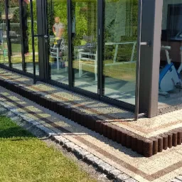 Wykończona ścieżka z kostki brukowej przed przeszklonym ogrodem zimowym z widocznym sprzętem do ćwiczeń wewnątrz.