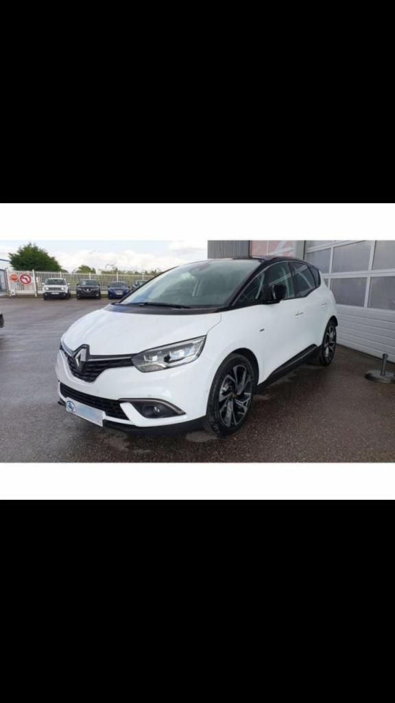 Biały samochód Renault Scenic z czarnym dachem i felgami, zaparkowany na zewnątrz przed budynkiem z bramą garażową.