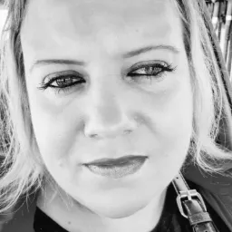 Czarno-biały portret kobiety z intensywnym spojrzeniem, widoczny fragment paska na ramieniu, oświetlenie z góry, podkreślone usta i rzęsy.