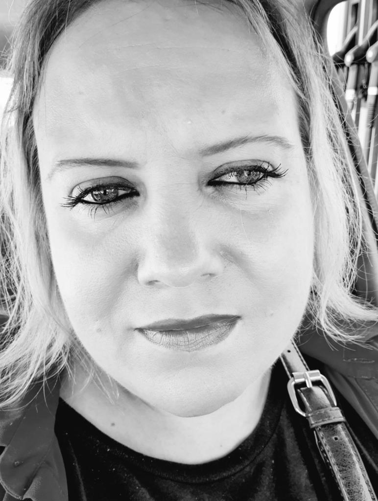 Czarno-biały portret kobiety z intensywnym spojrzeniem, widoczny fragment paska na ramieniu, oświetlenie z góry, podkreślone usta i rzęsy.