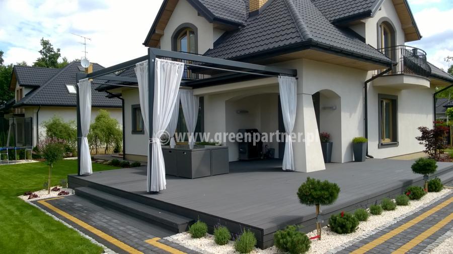 Zadaszony taras z szarej deski kompozytowej, osłonięty białymi zasłonami, przylegający do białego domu z ciemną dachówką. Widoczne schody, ozdobne kamienie, zadbany trawnik i krzewy.