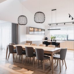 Wyskkuchnie - Jasna jadalnia z drewnianym stołem, szarymi krzesłami i nowoczesnymi lampami wiszącymi. W tle widoczna kuchnia z drewnianymi frontami i przeszklonymi szafkami.