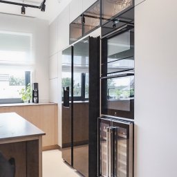 Wyskkuchnie - Nowoczesna kuchnia z zabudową w odcieniach bieli i drewna, czarny sprzęt AGD, oświetlenie szynowe i wyspa kuchenna z blatem w kolorze grafitowym.