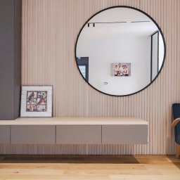 Wyskkuchnie - Nowoczesna aranżacja wnętrza: wisząca szafka z szufladami w kolorze taupe, okrągłe lustro i fotel na tle ściany z drewnianych paneli. Minimalizm i elegancja.