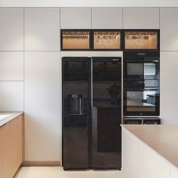 Wyskkuchnie - Nowoczesna kuchnia na wymiar w odcieniach bieli i beżu, z czarnym sprzętem AGD i podświetlaną witryną na kieliszki, minimalistyczny design.