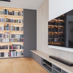 Wyskkuchnie - Nowoczesna szafa wnękowa z podświetlanymi półkami na książki, obok ściana z panelem dekoracyjnym i telewizorem. Jasna podłoga.