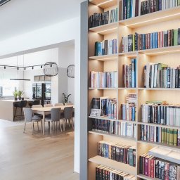 Wyskkuchnie - Regał na książki z oświetleniem LED, widoczny z perspektywy salonu z otwartą kuchnią. Jasne drewno i szare elementy wykończeniowe. Minimalistyczny design.