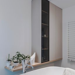 Wyskkuchnie - Jasna łazienka z wanną wolnostojącą, szafką wiszącą z przeszklonym frontem i drewnianą półką z roślinami doniczkowymi. Minimalistyczny design.