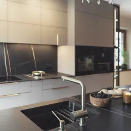 Nowoczesna kuchnia z czarnym blatem z żyłami, złotą armaturą i kryształowym żyrandolem. Minimalistyczne fronty szafek i oświetlenie LED dodają elegancji.