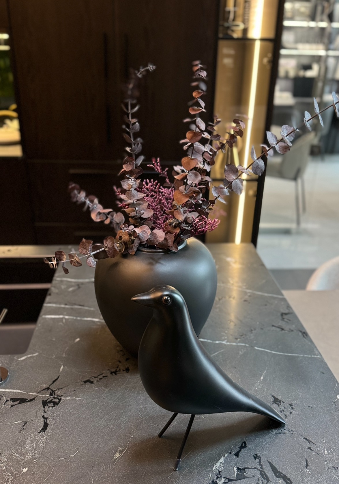 Czarny blat z marmuru, dekoracja z suszonych kwiatów w wazonie i figurka ptaka. Element wystroju kuchni, ciemne meble w tle.