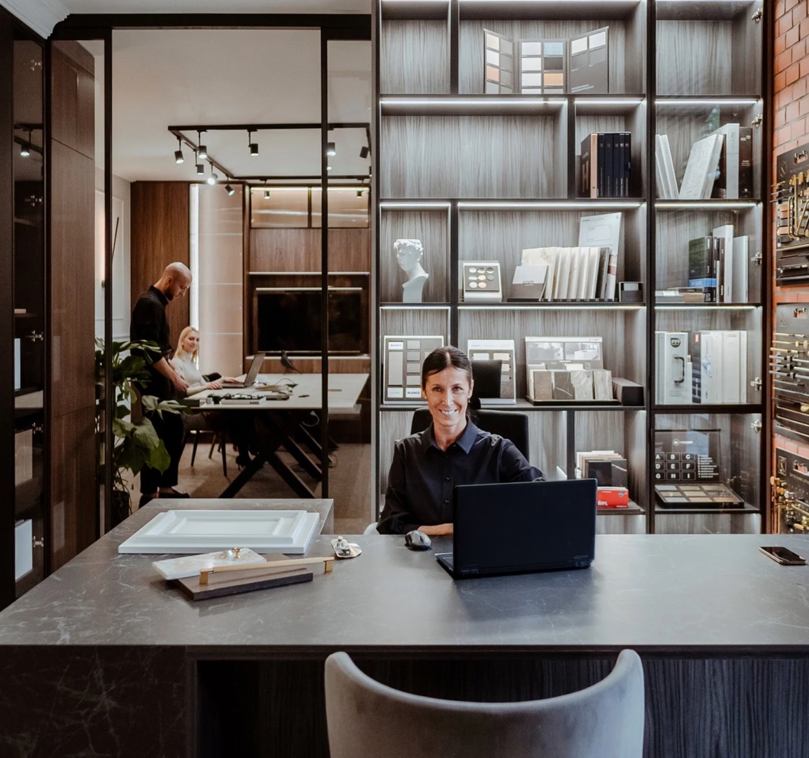 Kobieta przy biurku w salonie mebli na wymiar w Poznaniu. Na blacie próbki materiałów i laptop, w tle regał z ekspozycją frontów i blatów.