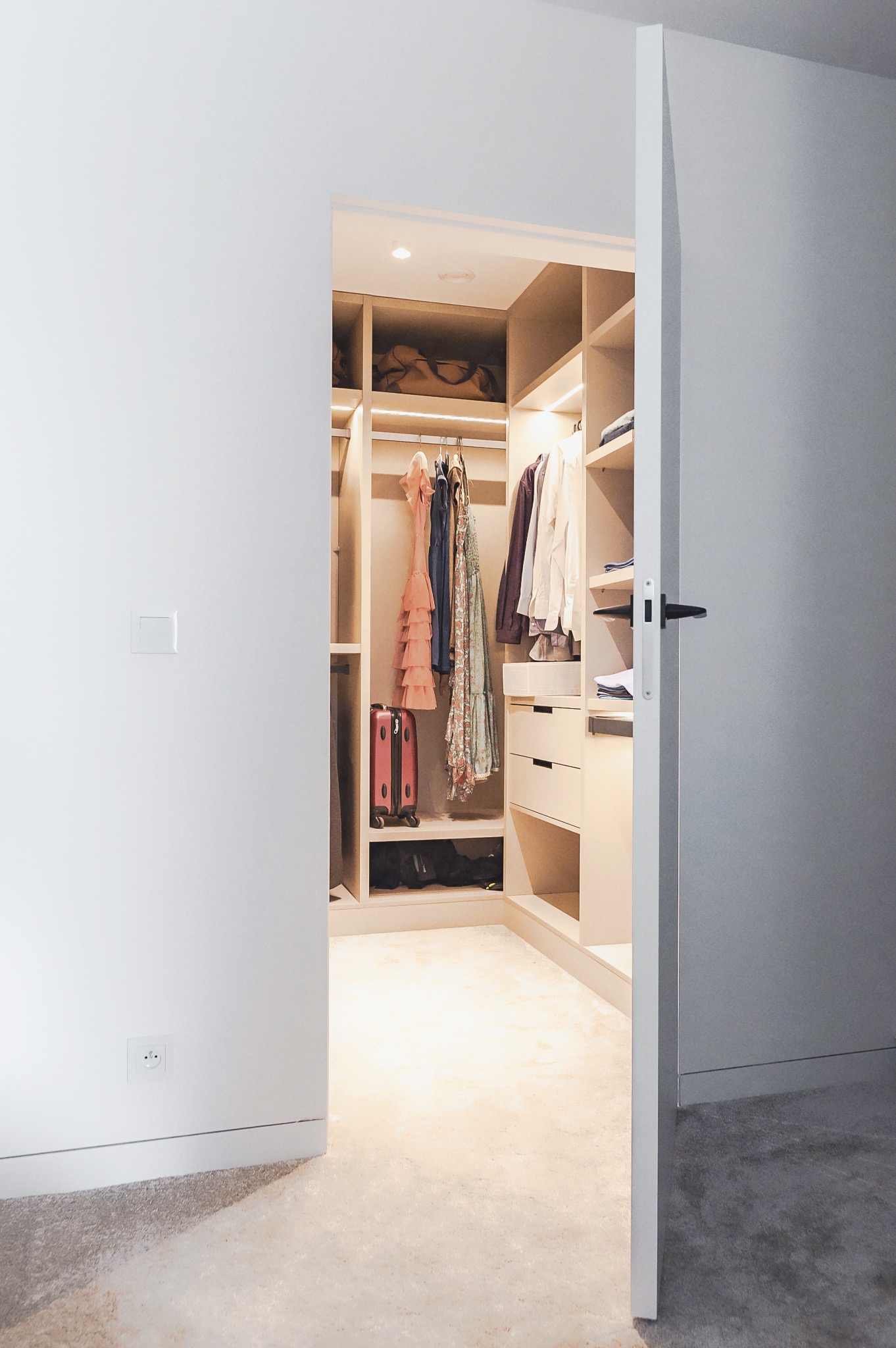 Otwarta szafa wnękowa z oświetleniem LED, ubraniami na wieszakach i walizką. Widok zza uchylonych drzwi, minimalistyczny design, jasne wnętrze.