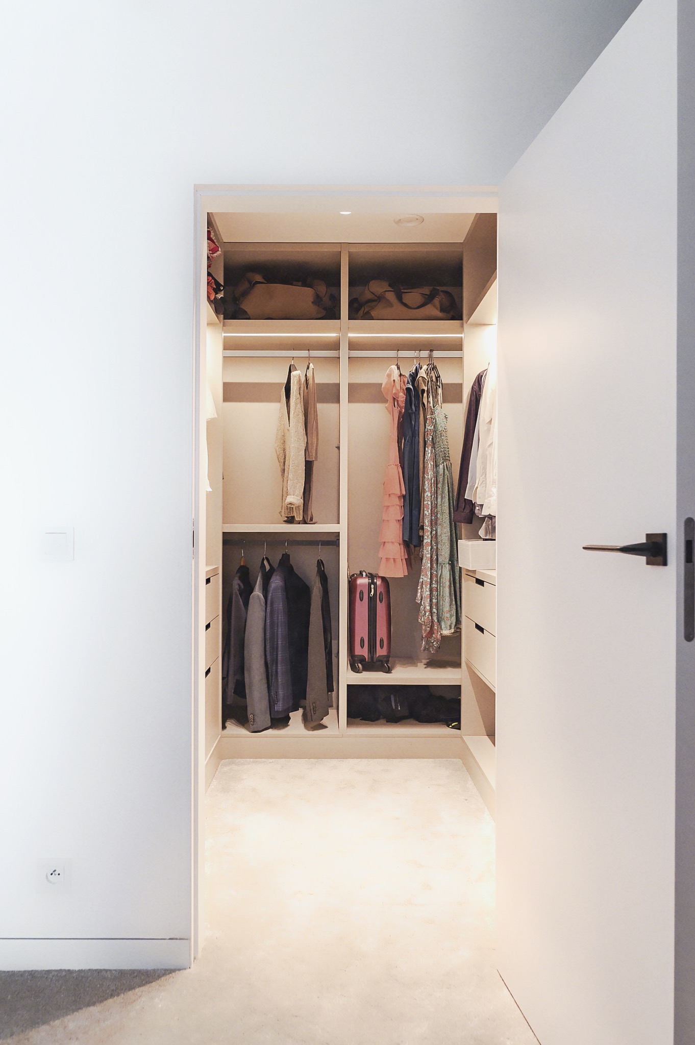 Otwarta, jasna garderoba wnękowa z ubraniami na wieszakach i półkach, widoczna z korytarza. Minimalistyczny design, ciepłe oświetlenie.