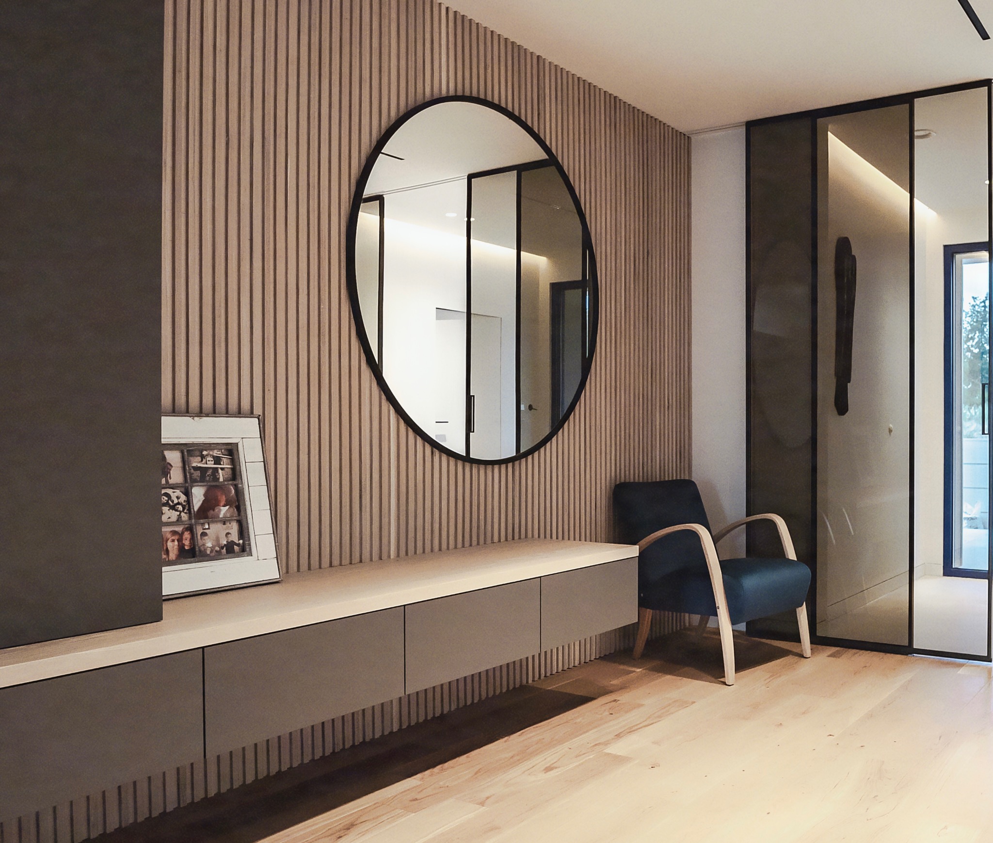 Nowoczesny przedpokój z szafką RTV, okrągłym lustrem i fototapetą z pionowymi lamelami. Na szafce ramka ze zdjęciami. Minimalistyczny design.